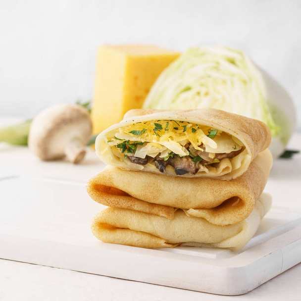 Stack of vegetarian crepes or blintzes with cabbage, mushrooms and cheese filling and ingredients for cooking. - Фото, изображение