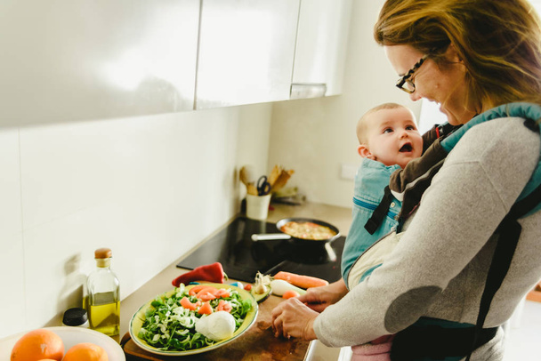 Bambino sorridente portato nello zaino del marsupio guardando sua madre mentre cucina, concetto di conciliazione familiare
 - Foto, immagini