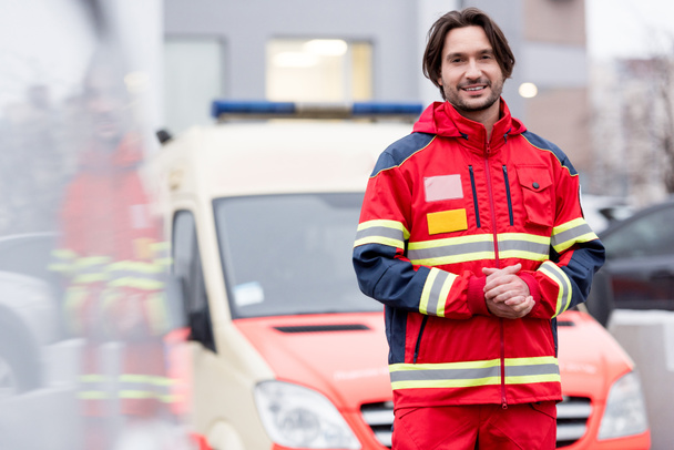 Sonriente paramédico en uniforme de pie cerca de coche ambulancia
 - Foto, Imagen