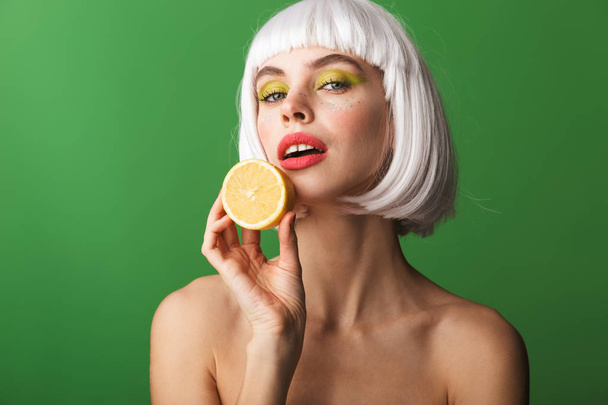 Attractive young topless woman wearing short white hair standing isolated over green background, showing sliced lemon - Fotó, kép