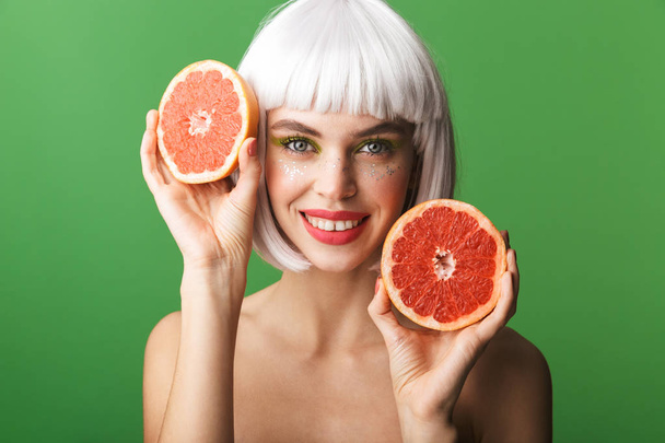 Beautiful healthy young topless woman wearing short white hair standing isolated over green background, showing sliced grapefruit - Foto, Imagen