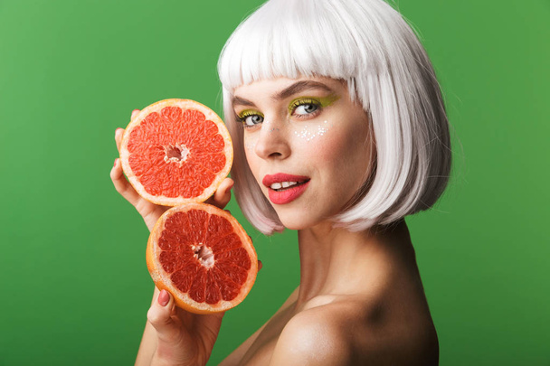 Beautiful healthy young topless woman wearing short white hair standing isolated over green background, showing sliced grapefruit - Фото, зображення