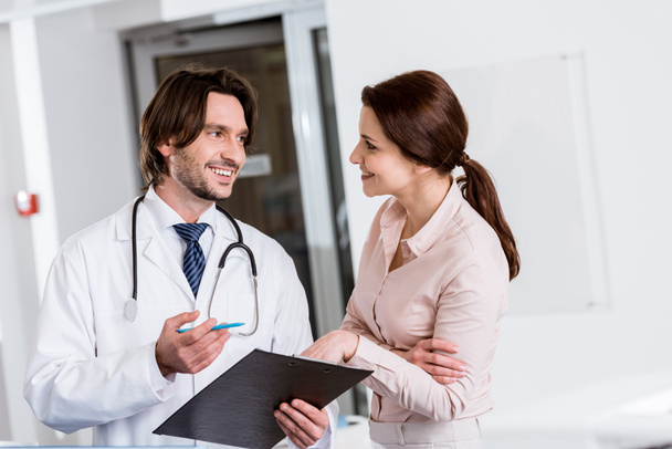 Handsome doctor holding clipboard and talking to patient in clinic - Foto, imagen