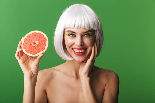 Beautiful healthy young topless woman wearing short white hair standing isolated over green background, showing sliced grapefruit - Φωτογραφία, εικόνα