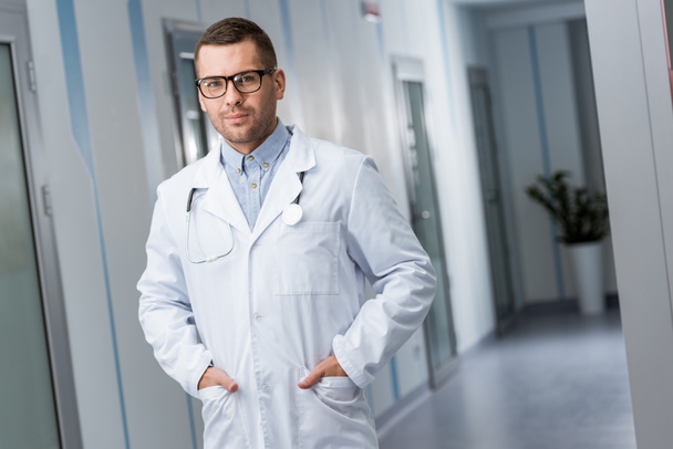 Doctor en gafas y abrigo blanco de pie con las manos en los bolsillos
 - Foto, Imagen