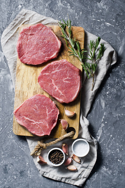 Solomillo de carne de res en una tabla de cortar madera, ajo y una ramita de romero. Vista superior, fondo gris
 - Foto, Imagen