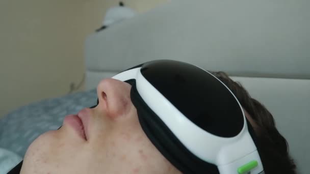 joven atractivo hombre utiliza un dispositivo masajeador de ojos, acostado en la cama, sosteniendo un control remoto, primer plano
 - Imágenes, Vídeo