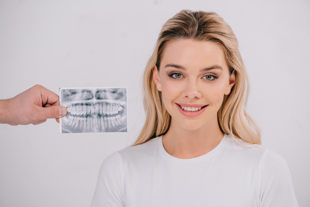 мужская рука держит рентген зубов в то время как красивая улыбающаяся женщина в футболке смотрит на камеру, изолированную на белом
 - Фото, изображение