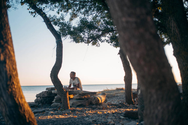 Um jovem senta-se junto ao mar ao pôr-do-sol. Um tipo bonito a olhar atenciosamente para o horizonte. Homem solitário encontra o pôr do sol em um pequeno bosque no oceano
 - Foto, Imagem
