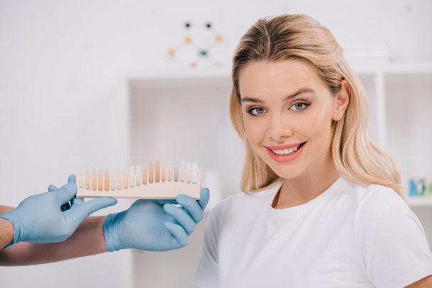dentista masculino sosteniendo los dientes paleta de colores mientras hermosa mujer sonriente mirando a la cámara en la clínica
 - Foto, Imagen