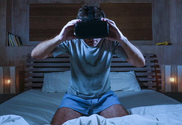 young excited and amazed man wearing VR virtual reality goggles device playing internet 3D simulator game enjoying exciting and shocking visual experience at home bed - Foto, afbeelding