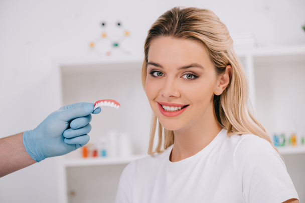 красивая улыбающаяся женщина смотрит в камеру, в то время как стоматолог в латексных перчатках держит покрытие зубов в клинике
 - Фото, изображение