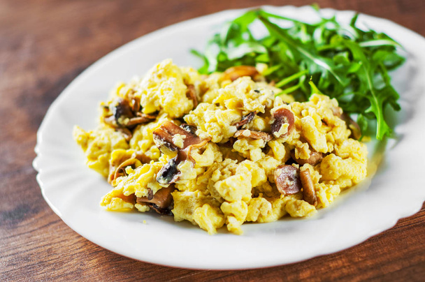 scrambled eggs with mushrooms and arugula salad in white plate on wooden table background - 写真・画像