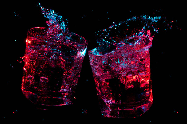 Grandes salpicaduras de líquido brillando en luces de colores en dos vasos de rocas tintineo en tostadas aisladas sobre un fondo negro
 - Foto, Imagen
