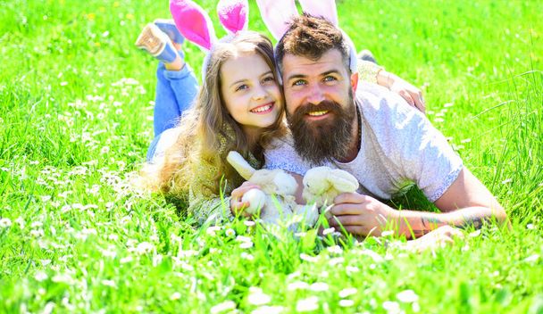 Father and child with cute bunny ears lying at meadow. Man with beard and cute child lay on grass with bunny toy in hands. Family feast concept. Dad and girl play in spring garden on sunny day - Foto, imagen