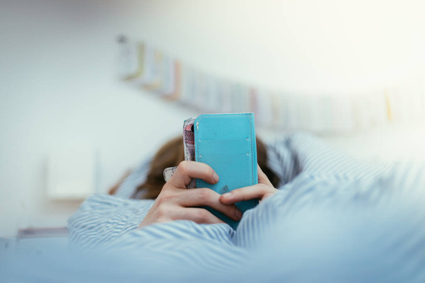 Girl lying in bed is using her smartphone, smartphone in the foreground, blurry background - Zdjęcie, obraz
