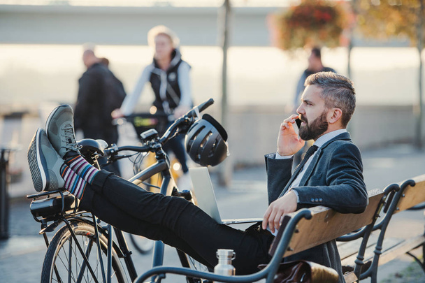 Businessman commuter with bicycle sitting on bench in city, making a phone call. - Foto, afbeelding