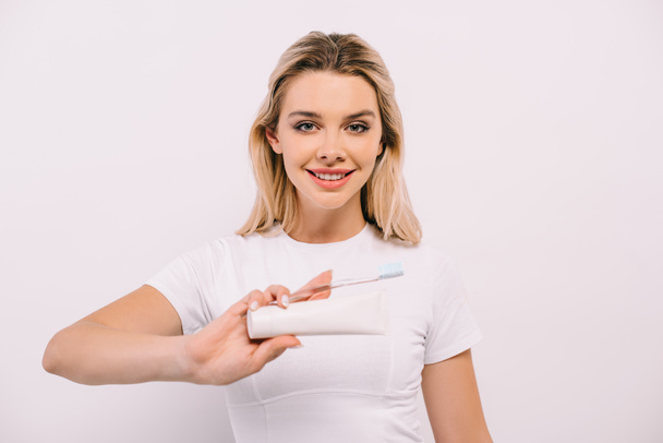 attractive smiling woman looking at camera while holding toothbrush and toothpaste with copy space isolated on white - 写真・画像