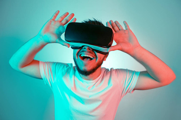 Photo of excited bearded man trying VR headset and exploring another world on the colorful background. Smartphone using with VR headset. - Фото, изображение