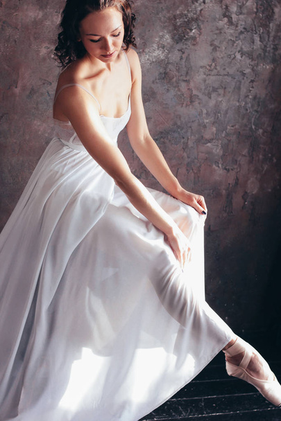 Ballet dancer ballerina in beautiful thin flying white dress is posing in dark loft studio - Фото, изображение