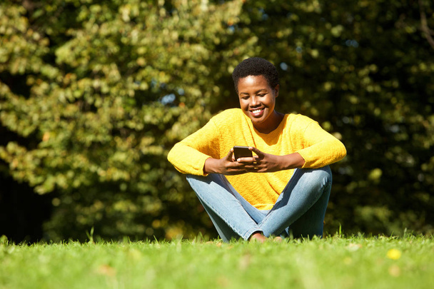 Porträt einer schönen jungen schwarzen Frau, die im Park sitzt und auf ihr Handy schaut  - Foto, Bild