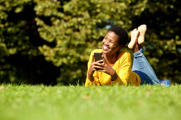 Portrait of happy young black woman lying in park with cellphone  - Fotoğraf, Görsel