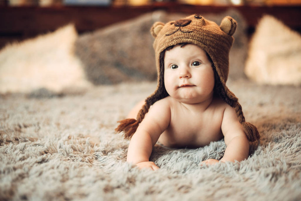 очаровательный ребенок 6 месяцев лежал на животе на кровати в кепке медведя в интерьере
 - Фото, изображение