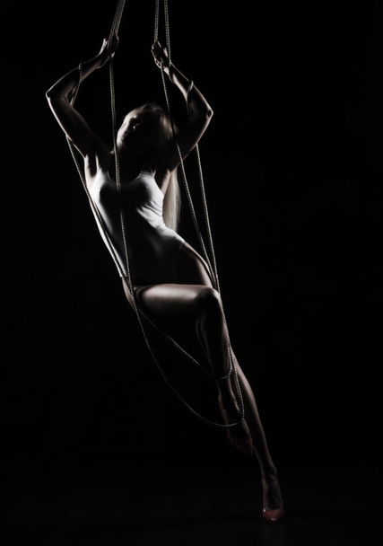 Beautiful slender blonde girl, wearing a white bodisuit and red stilettos, sensually hangs and dances on the ropes. Black background. Artistic noir silhouette photo. Copy space. - Foto, Imagem