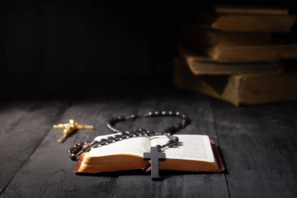 Open book of Bible and crucifix on dark table. Low-key image of New Testament, cross and rosary in bright light among darkness and shadows with copy space - Foto, Imagem
