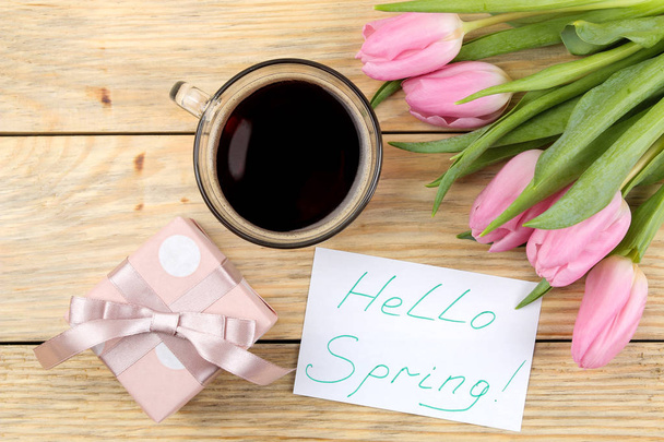 Beautiful bouquet of pink tulips flowers, cup of coffee, gift box and text hello spring on paper on a natural wooden background. view from above. Spring. holidays. - Фото, изображение