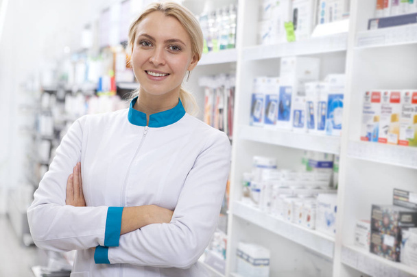 Cheerful confident female pharmacist smiling to the camera with her arms crossed, posing proudly at her drugstore. Attractive female chemist working at the pharmacy, copy space. Occupation, career concept - Photo, Image