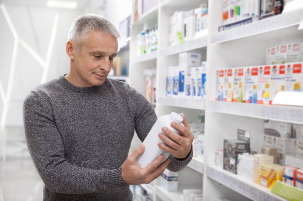 Happy mature man buying medications at the local pharmacy, copy space. Cheerful senior man examining bottle of a medical product, while shopping at drugstore. Health, quality, retail concept - Photo, Image