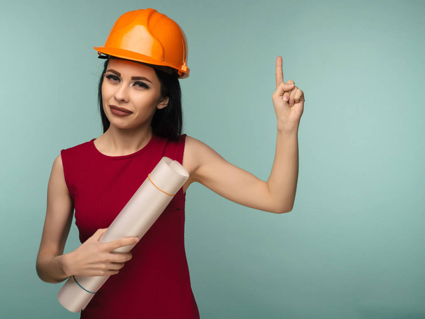 Молодая женщина-инженер в оранжевом шлеме с рисунками указывает пальцем вверх на синий фон - Изображение
 - Фото, изображение