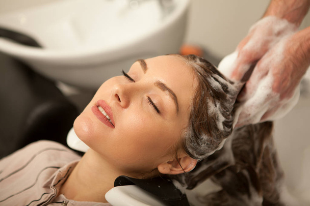 Close up of a beautiful young woman enjoying head massage with her eyes closed, while professional hairdresser washing her hair with shampoo. Hairstylist working with his female client - Foto, afbeelding
