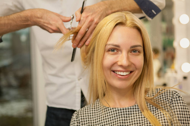 Charming blond haired woman smiling to the camera while receiving new haircut, copy space. Happy young woman enjoying getting new hairstyle at beauty salon, copy space, Retail, service concept - Zdjęcie, obraz