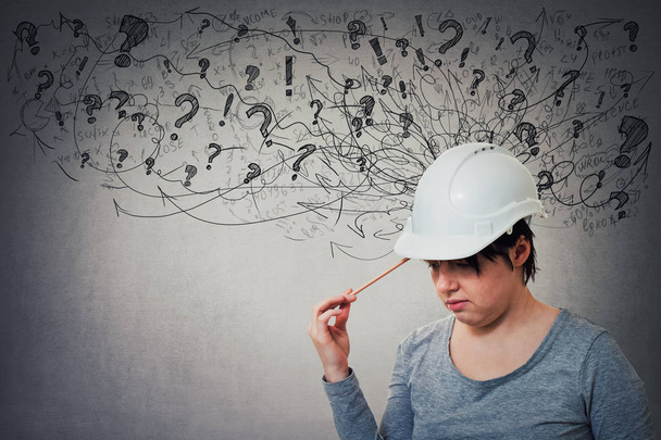 Thoughtful young woman engineer wearing protective helmet thinking pointing pencil to head, stressed looking down feeling exhausted. Headache, anxiety and health problems as sketch mess in his head. - Photo, Image