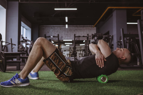 Full length shot of a male athlete using foam roller after working out at the gym. Sportsman massaging and stretching his back muscles with foam roller. Health, relief, injury concept - Фото, изображение
