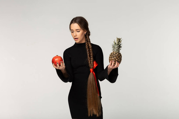 Model with long braid and red bow in black dress can not choose what eat first, pineapple or garnet - Photo, Image