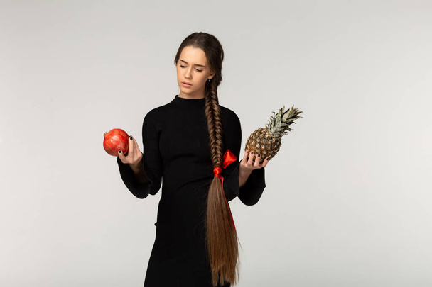 Model met lange vlecht en rode strik in zwarte jurk kan niet kiezen wat eet eerst, ananas of garnet - Foto, afbeelding
