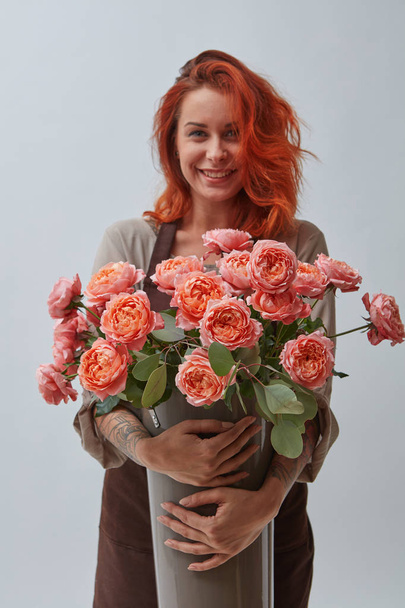 A smiling red-haired woman in a brown apron holds a vase with roses in a color of the year 2019 Living Coral Pantone on a gray background with space for text. The concept of a flower shop - Фото, изображение