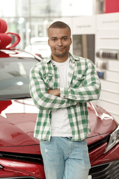 Vertical shot of a young handsome African man posing proudly in front of his new automobile at the dealership salon. Happy male driver leaning on his new car. Consumerism, rental service concept - Photo, Image