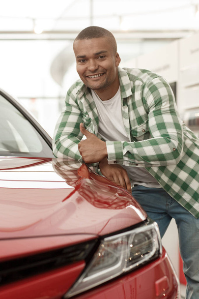 Vertical portrait of a happy handsome African man smiling, showing thumbs up, leaning on his new automobile. Cheerful male driver posing near new car. Buying cars, car rental service, travel concept - Photo, Image