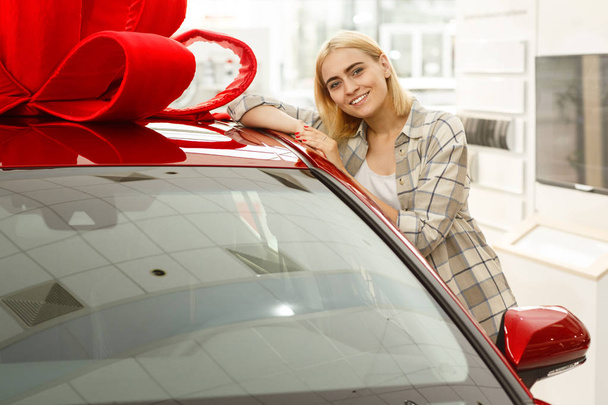 Attractive young happy woman smiling cheerfully to the camera leaning on her new automobile at the dealership, copy space. Beautiful female customer buying new car. Woman receiving auto as present - Valokuva, kuva