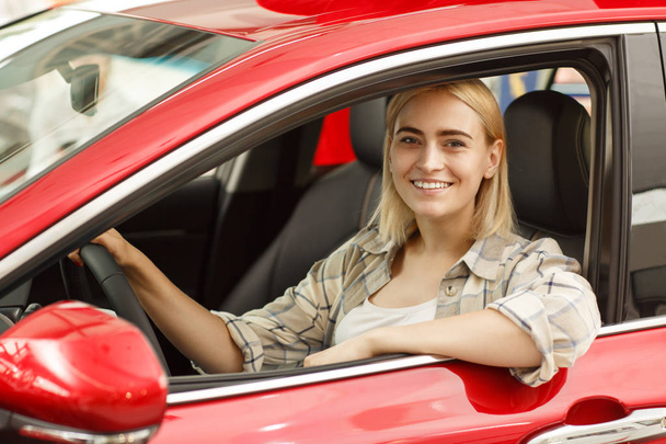 Piękny szczęśliwy blond kobieta uśmiecha się radośnie do kamery, siedząc w nowy samochód w salonie dealera. Atrakcyjna kobieta-kierowca ciesząc się jej nowy samochód, siedzący spokojny, kierownicy gospodarstwa - Zdjęcie, obraz