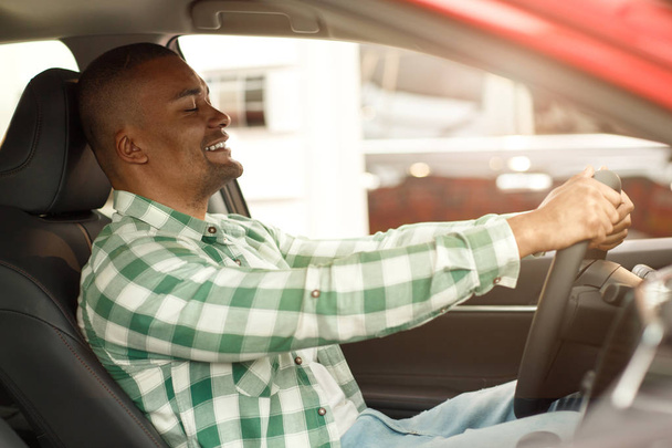 Foto de un hombre africano guapo sonriendo con los ojos cerrados disfrutando sentado en un nuevo automóvil cómodo en el concesionario. Cliente masculino que elige el coche para comprar. Estilo de vida, conducción, concepto de seguridad
 - Foto, imagen