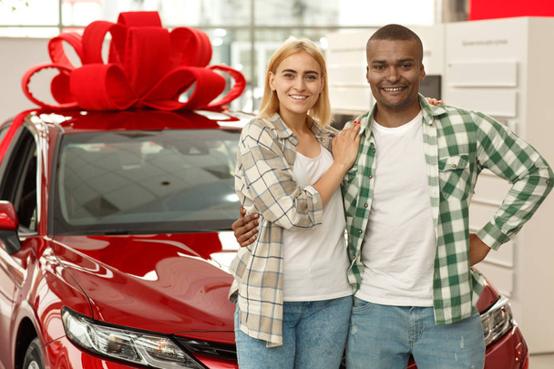Horizontal shot of a happy young interracial couple embracing smiling to the camera standing in front of their new automobile at the dealership salon, copy space. Anniversary, gift, winning concept - 写真・画像
