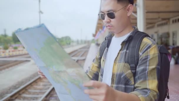 Utazó ázsiai ember irányát és látszó-on Térkép-míg a kiadások nyaralás utazás és a vasútállomáson várakozó vonat, hátizsákos fiatal férfi turista élvez a utazás. Életmód-férfi utazási koncepció. - Felvétel, videó