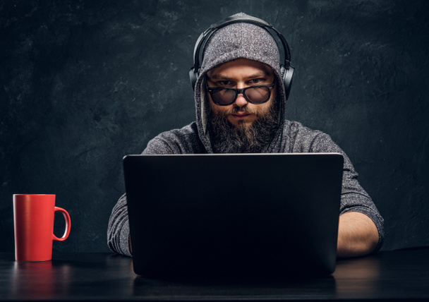 Ένας χάκερ κάθεται πίσω από ένα φορητό υπολογιστή σε ένα γραφείο σκούρο και κοιτάζει επίμονα με προσήλωση στην κάμερα - Φωτογραφία, εικόνα