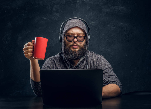 Хакер сидит за ноутбуком, держа чашку кофе и работая с программами в темной комнате
 - Фото, изображение