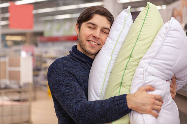 Happy handsome young man smiling with his eyes closed, embracing pile of soft new pillows he is buying at furniture store. Cheerful man shopping for beddings at home goods market, copy space - Photo, image